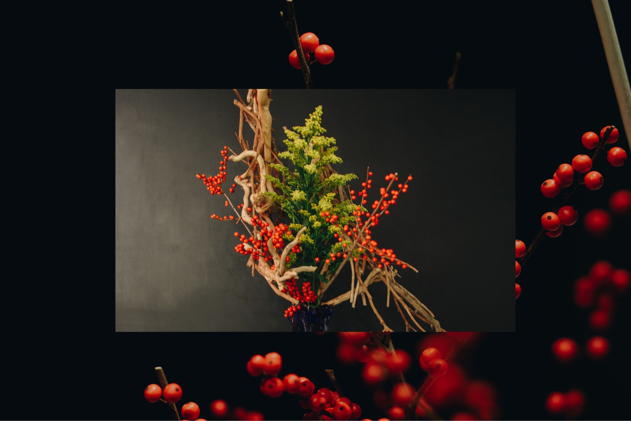 ikebana floral artist work of red buds, wood, and greenery in a vase against a black background