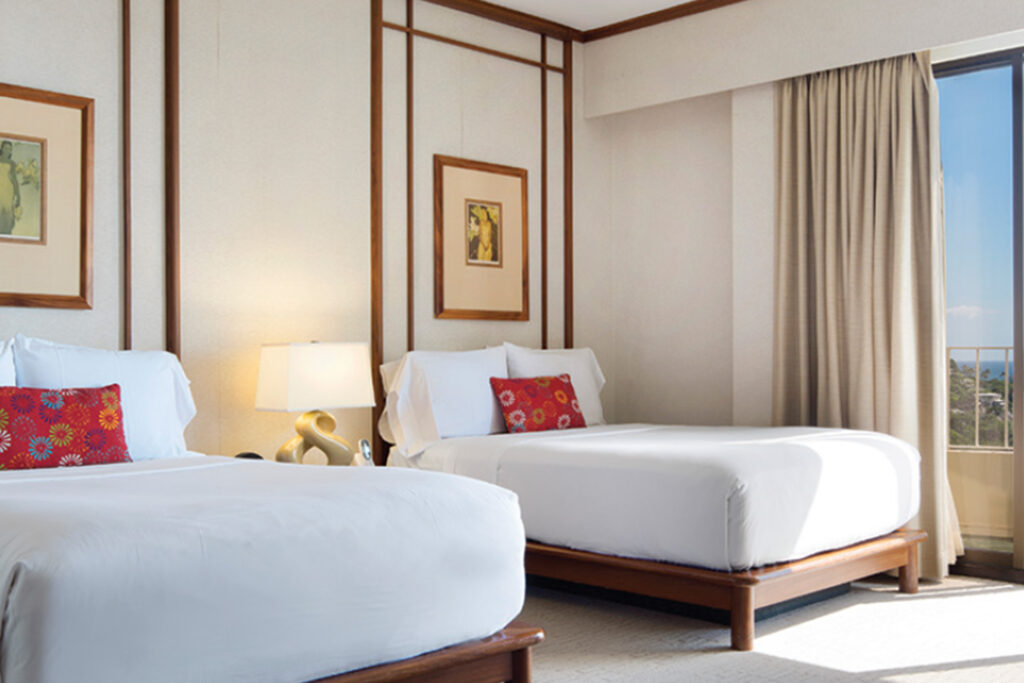 Hotel room with floor to ceiling windows. Inside the room there are beautiful dark wood, upholstered furniture, making it really feel fresh and bright like the beach