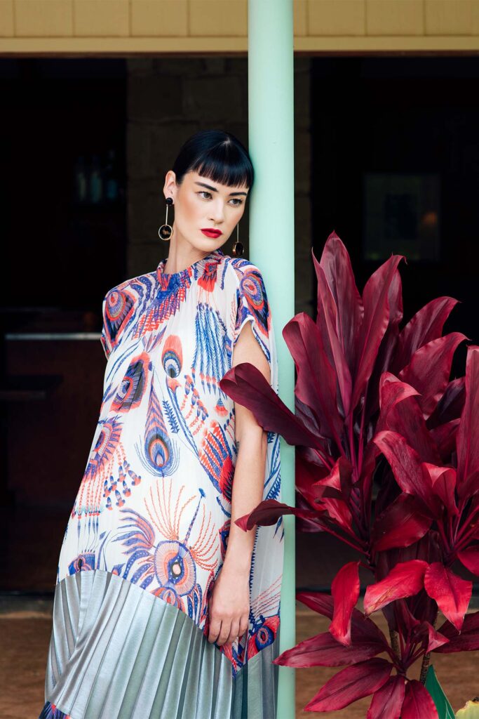multicolored pleated dress, worn by a model with a bold red lip. Model leans against a pole surrounded by red leafed plants. 