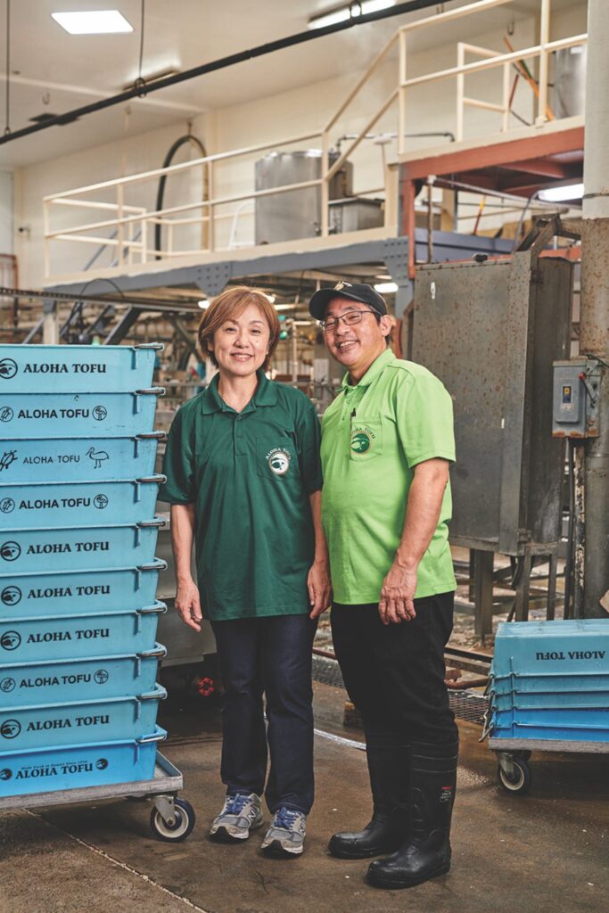 Aloha tofu owners stand in the factory next to blue crates